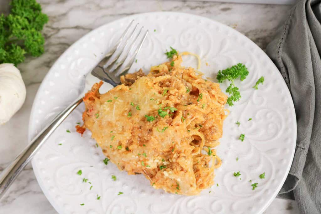 A serving of Spaghetti Squash Bake with melted cheese and ground meat, garnished with parsley, on a white plate with a fork.
