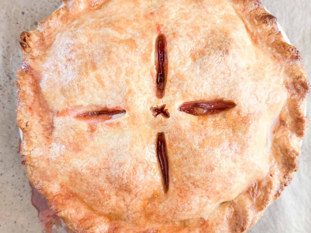 Golden brown strawberry rhubarb pie with four vents cut into the top crust, arranged in a cross pattern, resting on a light-colored surface.