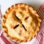A baked strawberry rhubarb pie with a golden crust and four slits in the center sits on a white surface between two red and white striped cloth napkins. A metal pie server is beside it.