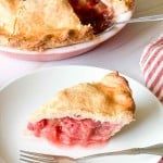 A slice of strawberry rhubarb pie on a white plate with a fork, with the remaining pie and a red-striped napkin in the background.