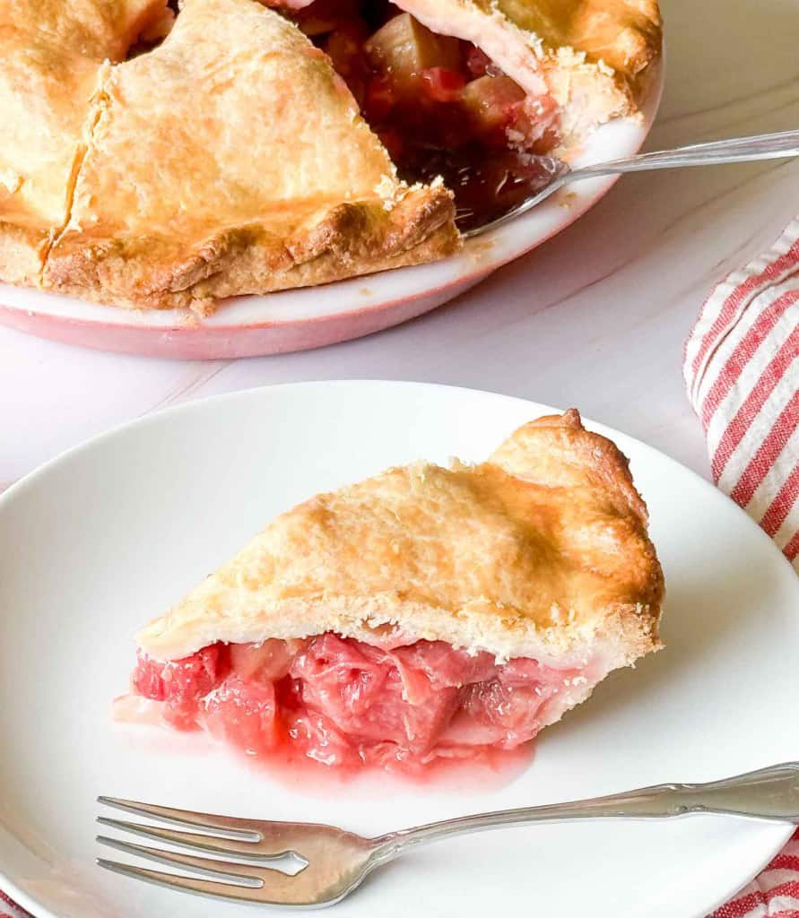 A slice of strawberry rhubarb pie on a white plate with a fork, with the remaining pie and a red-striped napkin in the background.