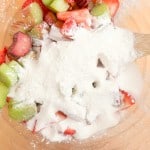Chopped rhubarb and strawberries in a glass bowl are topped with white sugar and flour, being stirred with a wooden spoon&mdash;the perfect filling for a classic strawberry rhubarb pie.