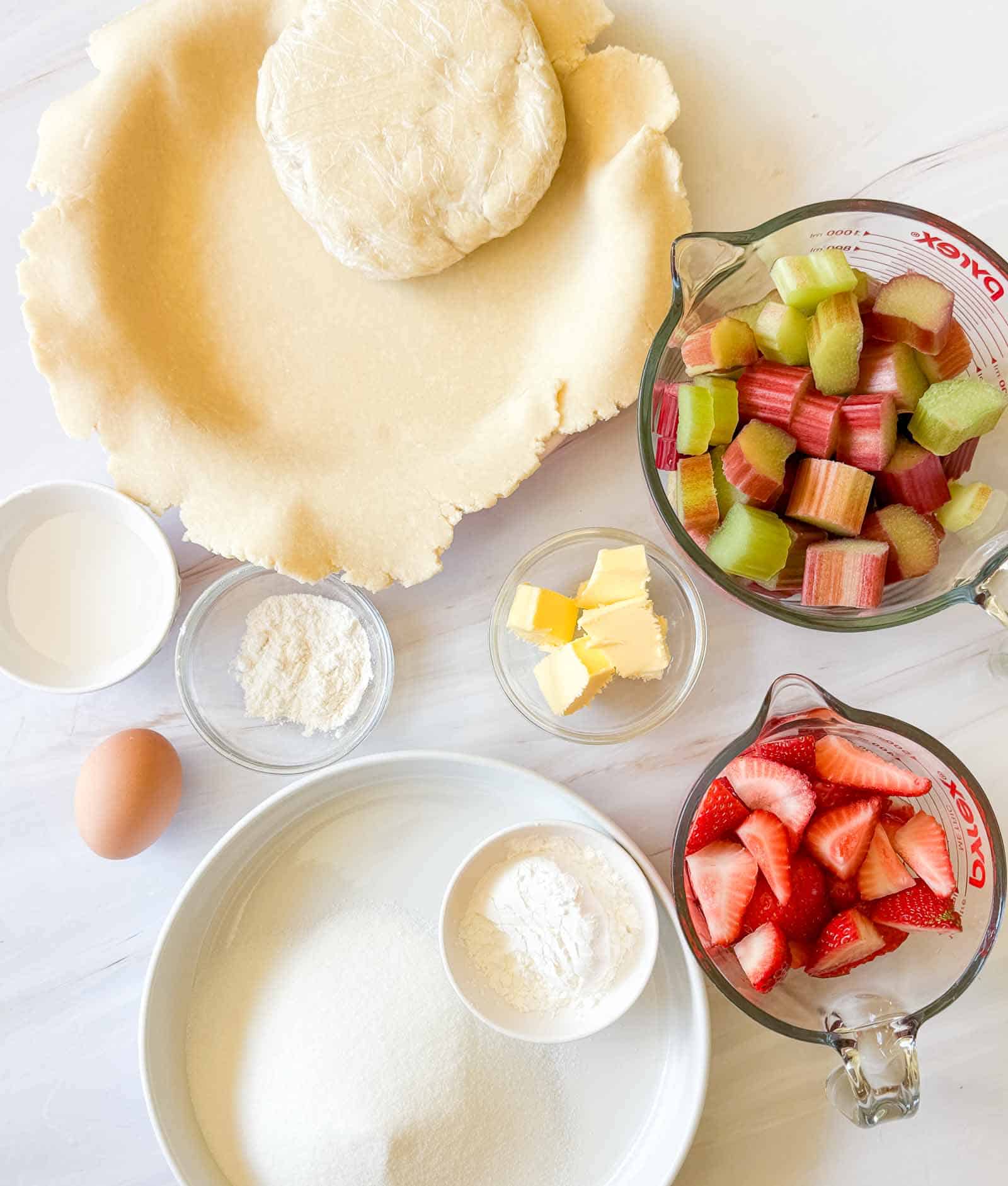 Pie dough, rolled crust, cubed butter, sugar, flour, egg, milk, chopped rhubarb, and sliced strawberries ready in bowls&mdash;the classic beginnings for a homemade strawberry rhubarb pie displayed on a white surface.