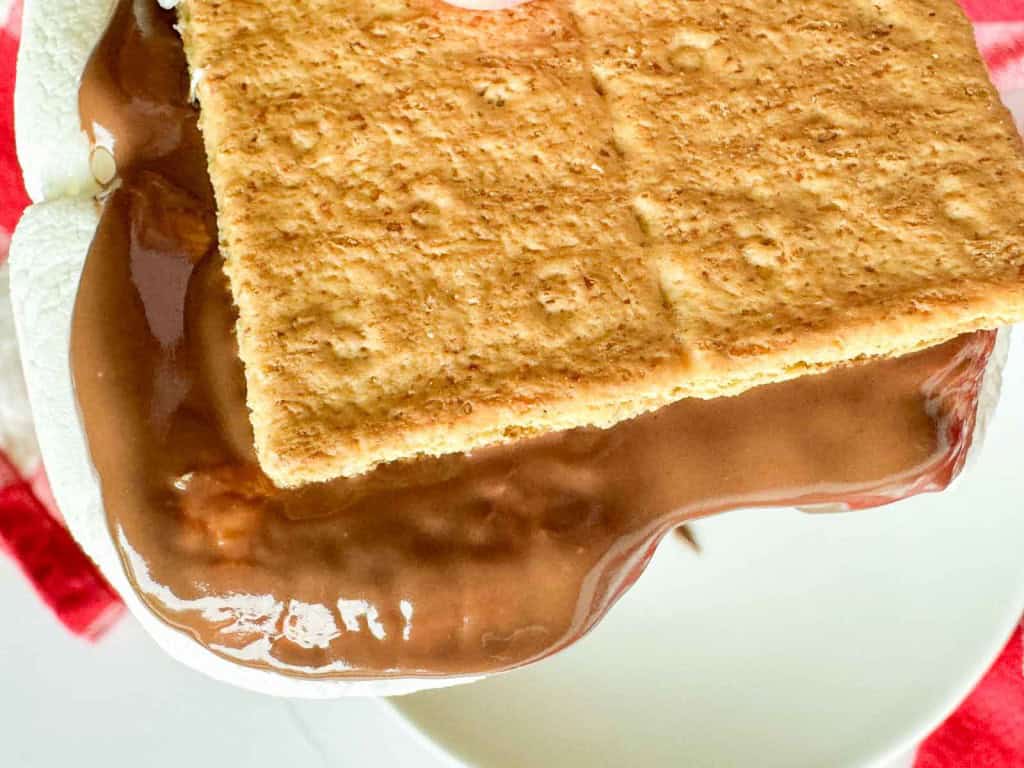 Close-up of a s&rsquo;more with melted chocolate and marshmallow between two graham crackers, resting on a white surface.