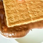 Close-up of a s&rsquo;more with melted chocolate and marshmallow between two graham crackers, resting on a white surface.