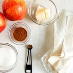 Two apples, a bowl of butter, phyllo dough sheets, sugar, ground cinnamon, and a measuring spoon of spice arranged on a white surface.