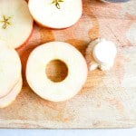 Sliced apples with the cores removed are arranged on a wooden cutting board. A small pile of apple cores and a container of ground cinnamon are nearby.