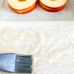 A pastry brush applies melted butter to a sheet of phyllo dough. Apple slices and a bowl of liquid are in the background.