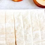 Rectangular strips of uncooked pastry dough are laid out on a white surface, with slices of cored apple in the background.