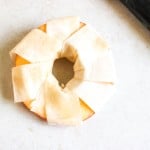 A round apple slice with a hole in the center, wrapped in overlapping strips of pastry dough, sits on a light surface next to a black-handled utensil.