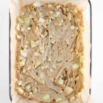 A glass baking dish lined with parchment paper containing unbaked blondie batter with visible white chocolate chips and apple chunks.