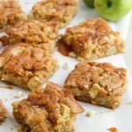 Nine caramel apple blondies on parchment paper, with two green apples in the background. Some blondies have a caramel drizzle and a few white chocolate chips are visible on the surface.