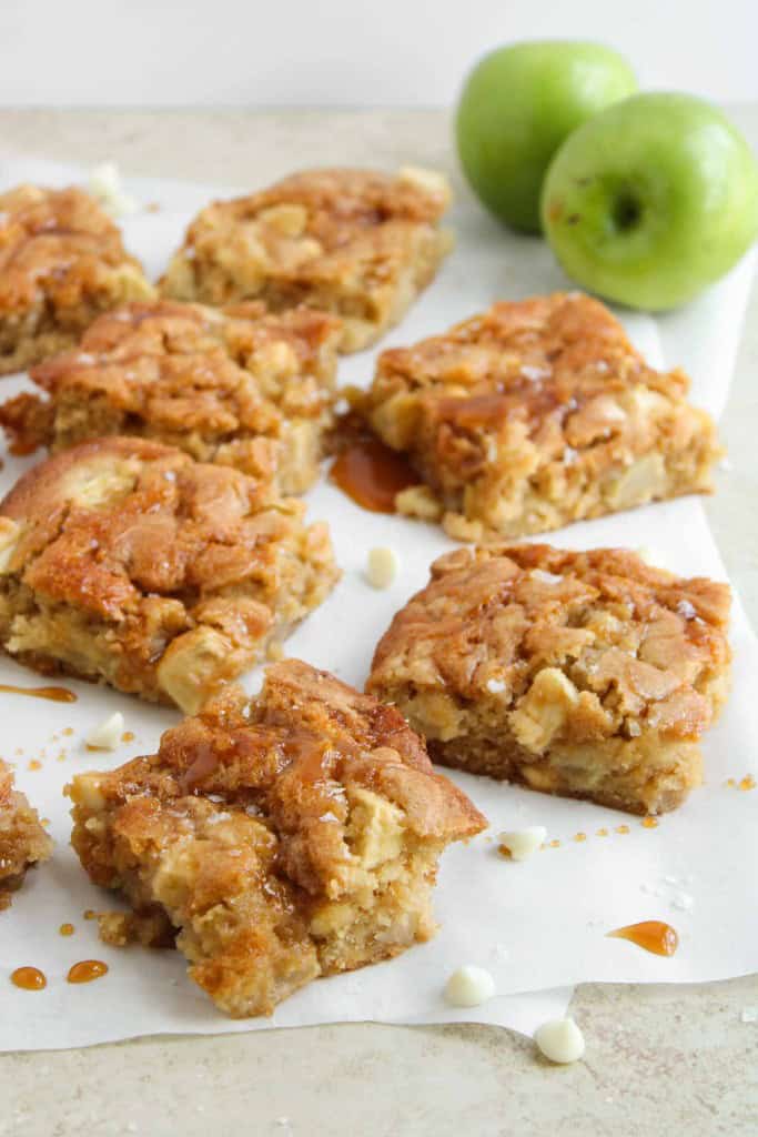 Nine caramel apple blondies on parchment paper, with two green apples in the background. Some blondies have a caramel drizzle and a few white chocolate chips are visible on the surface.