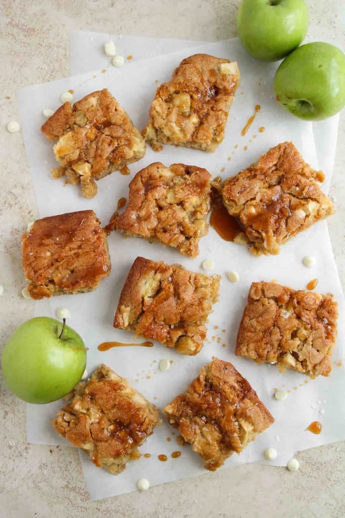 Nine caramel-drizzled apple blondie squares are arranged on parchment paper, with three green apples and some scattered white chocolate chips nearby.