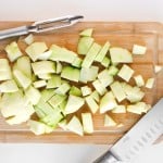 A chopped green apple on a wooden cutting board with a peeler and a knife placed nearby.