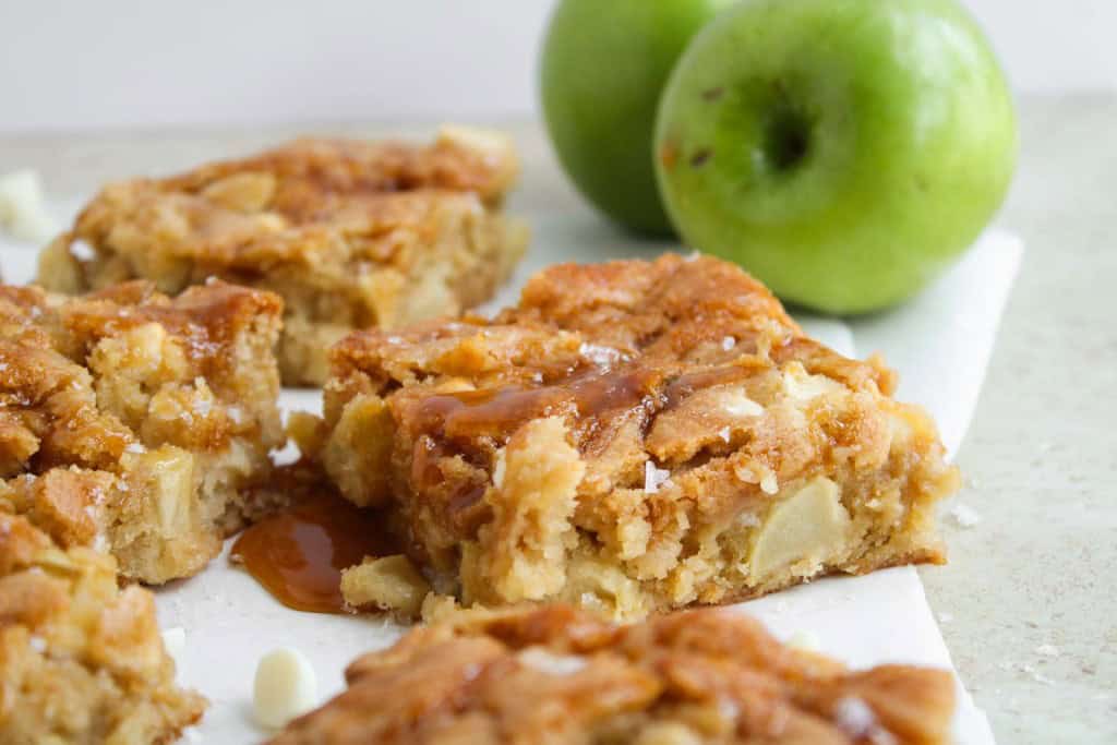 Apple blondies with caramel drizzle are arranged on a white surface, with two whole green apples in the background.