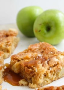 A square piece of apple cake drizzled with caramel sauce sits on a plate, with two whole green apples in the background.