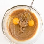 A glass bowl with brown batter, two raw eggs, and a metal whisk on a white surface.