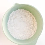 A green mixing bowl filled with white flour is seen from above on a white background.