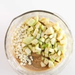 A glass bowl containing chopped green apples, white chocolate chips, and batter on a white surface.
