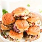 A white plate holds a stack of jalape&ntilde;o popper sliders with beef patties, red onions, bacon, cheese, and sauce on toasted buns.