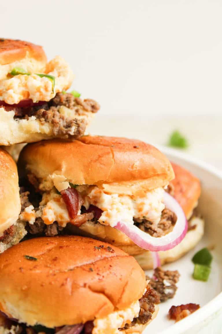 Three cheeseburgers topped with ground beef, creamy cheese spread, bacon pieces, red onion slices, and a hint of spice evoke the bold flavors of jalape&ntilde;o popper sliders as they are stacked on a white plate.