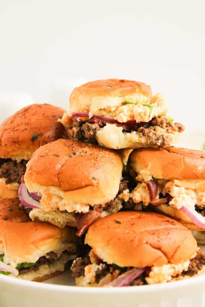 A stack of jalape&ntilde;o popper sliders with ground beef, lettuce, red onion, and sauce on toasted buns, arranged on a white plate.