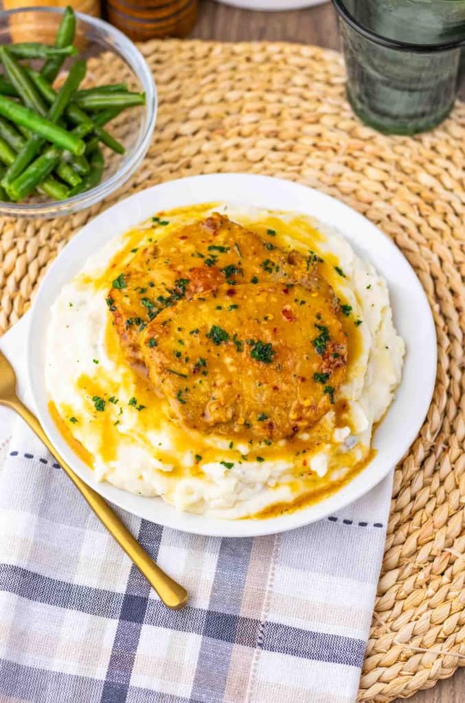 A plate of creamy mashed potatoes topped with a maple dijon pork chop in savory gravy, garnished with fresh herbs, sits on a woven placemat next to a bowl of green beans and a gold-colored fork.