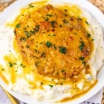 Two maple dijon pork chops in brown gravy are served on a mound of mashed potatoes, garnished with parsley, on a white plate with a woven placemat underneath.
