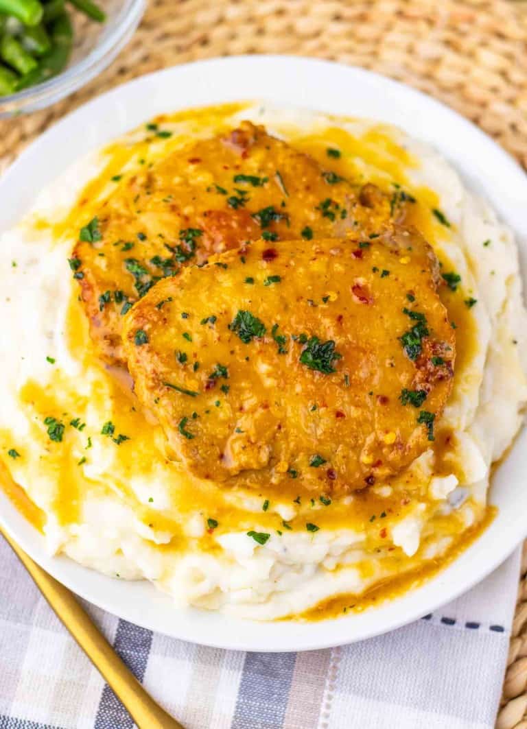 Two maple dijon pork chops in brown gravy are served on a mound of mashed potatoes, garnished with parsley, on a white plate with a woven placemat underneath.
