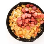 A skillet filled with sliced sausage, potatoes, cabbage, and carrots on a white background.