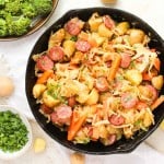 A skillet filled with sliced sausage, potatoes, carrots, and cabbage, garnished with parsley. Broccoli and chopped herbs are in bowls beside it.
