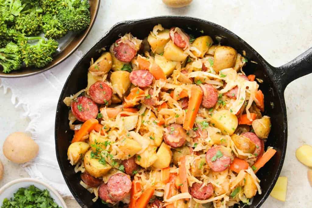 A cast iron skillet filled with sliced sausages, potatoes, carrots, cabbage, and herbs, with a side of steamed broccoli on the table.