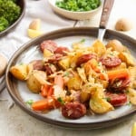 A plate with roasted potatoes, sliced sausage, carrots, cabbage, and herbs. Small potatoes, chopped parsley, and broccoli are visible in the background.