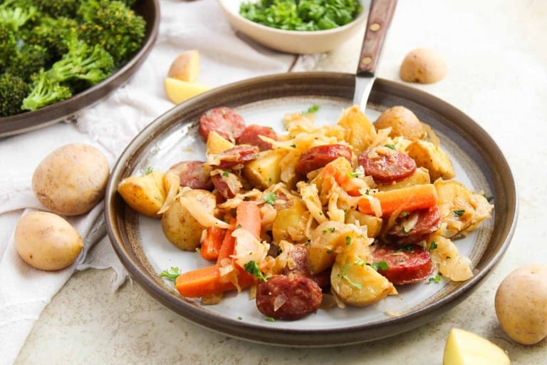 A plate with roasted potatoes, sliced sausage, carrots, cabbage, and herbs. Small potatoes, chopped parsley, and broccoli are visible in the background.