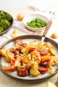 A plate of cooked potatoes, carrots, sausage slices, and onions garnished with parsley, with broccoli and chopped herbs in the background.