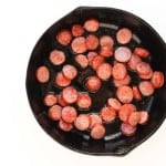 Sliced sausage pieces frying in a black cast iron skillet with some oil, viewed from above on a white background.