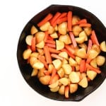 A black cast iron skillet filled with chopped potatoes and carrots, ready to be cooked, photographed on a white background.
