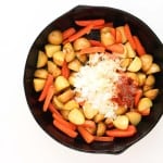 A black skillet with chopped baby potatoes, carrot sticks, diced onions, and seasonings on a white background.