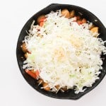 A black skillet filled with chopped vegetables and topped with shredded cabbage and pepper, photographed on a white background.