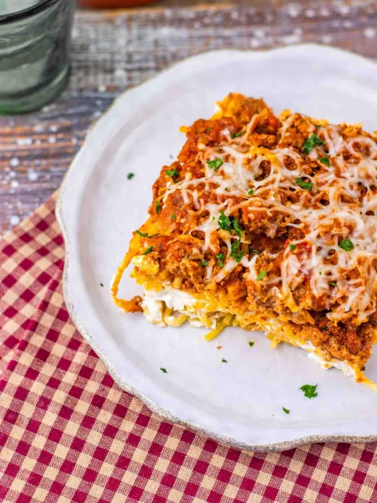 A slice of lasagna topped with melted cheese and herbs is served on a white plate, placed on a red and white checkered napkin, reminiscent of classic Million Dollar Spaghetti comfort.