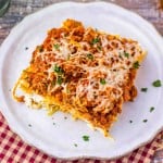 A square slice of Million Dollar Spaghetti lasagna topped with melted cheese and parsley is served on a white plate, set on a rustic wooden table with a red checkered cloth.