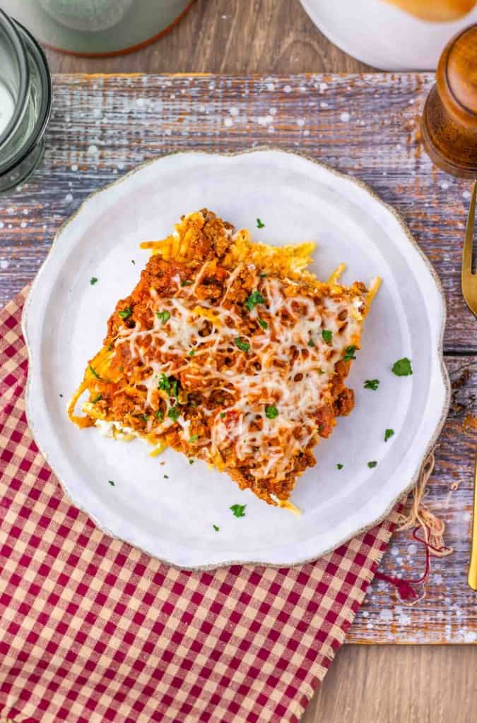 A square serving of Million Dollar Spaghetti baked pasta, topped with melted cheese and herbs, sits on a white plate placed on a red checkered cloth and wooden table.
