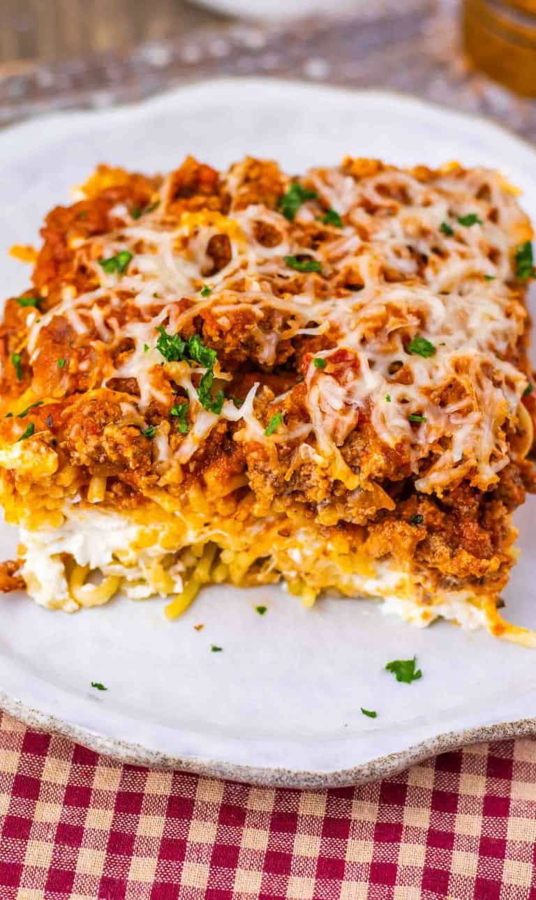 A slice of lasagna topped with melted cheese and parsley sits on a white plate, showing layers of pasta, meat sauce, and cheese&mdash;reminiscent of the rich, comforting flavors found in Million Dollar Spaghetti.