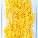 Uncooked yellow spaghetti noodles, ready for a delicious Million Dollar Spaghetti recipe, are arranged in a clear glass baking dish on a white background.