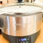 A stainless steel Farberware slow cooker sits on a kitchen counter with its digital display showing 23:50. Utensils are visible in the background.