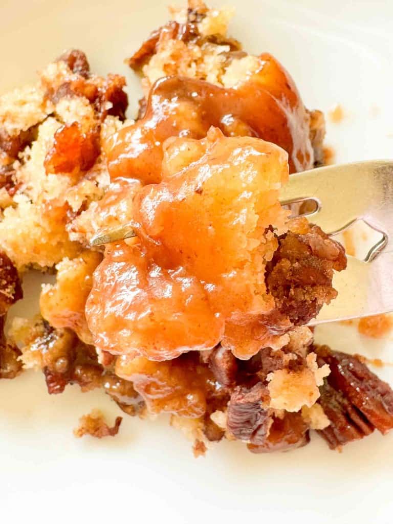 A close-up of a fork holding a bite of peach cobbler with pecans on a white plate.