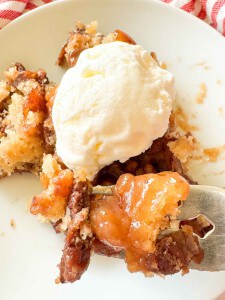 A serving of peach cobbler topped with a scoop of vanilla ice cream on a white plate, with a fork holding a bite-sized piece.