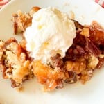 A serving of peach cobbler topped with a scoop of vanilla ice cream on a white plate, placed on a red and white striped cloth.