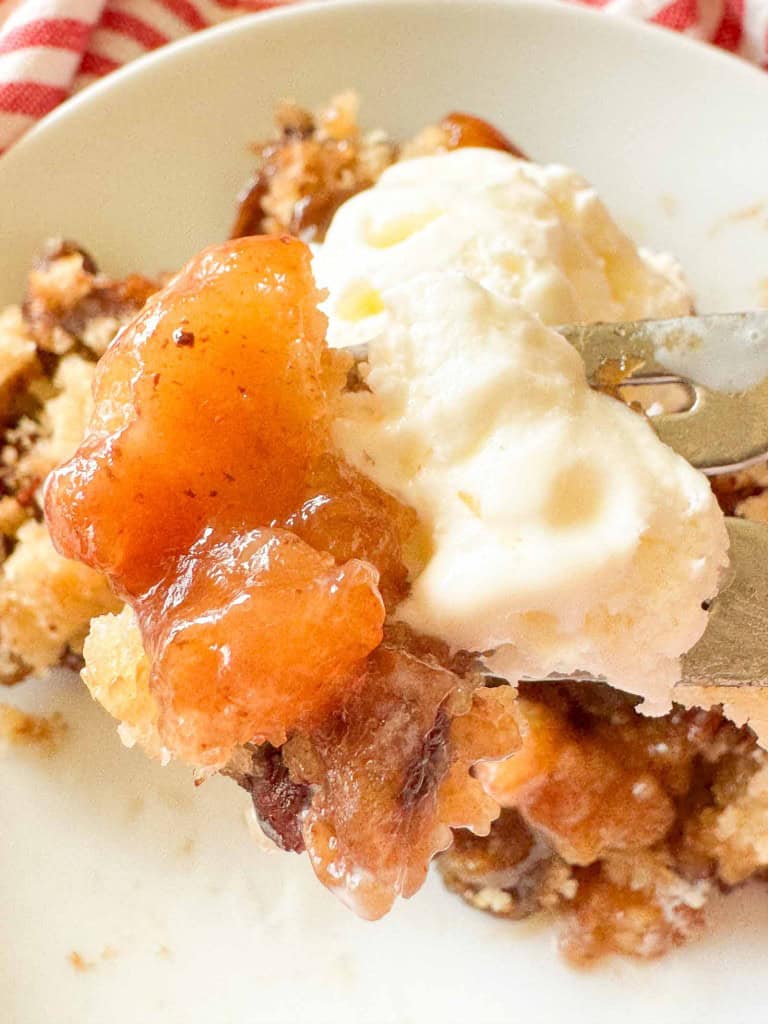 A fork holds a bite of apple cobbler topped with vanilla ice cream on a white plate.